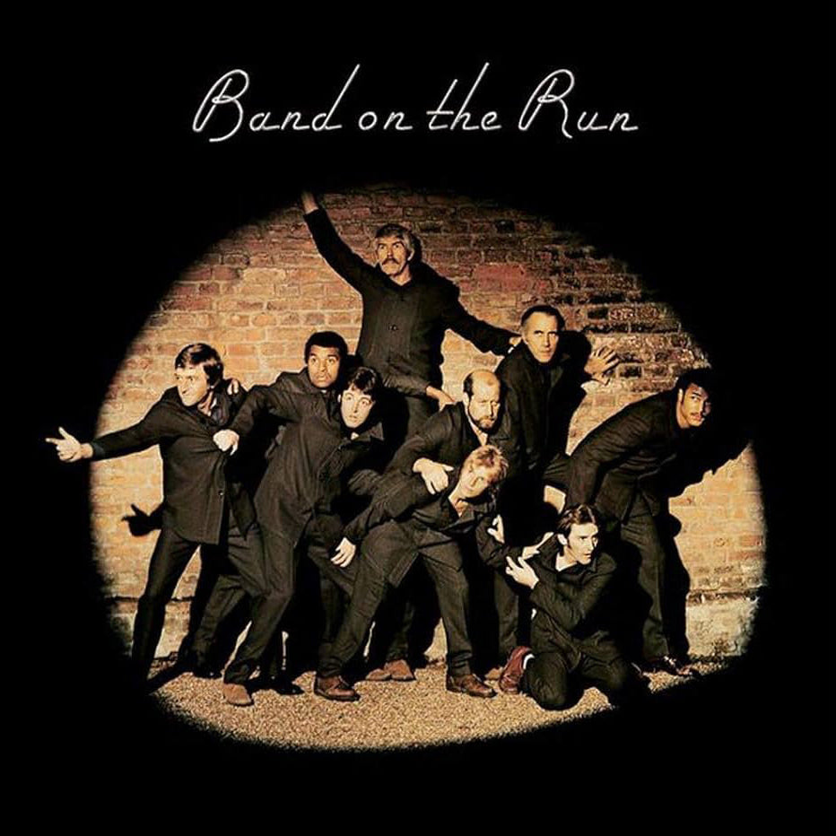 Album cover for 'Band on the Run' featuring a group of seven men posing dynamically inside a circular spotlight effect against a brick wall, set on a solid black background with the album title in white script at the top.
