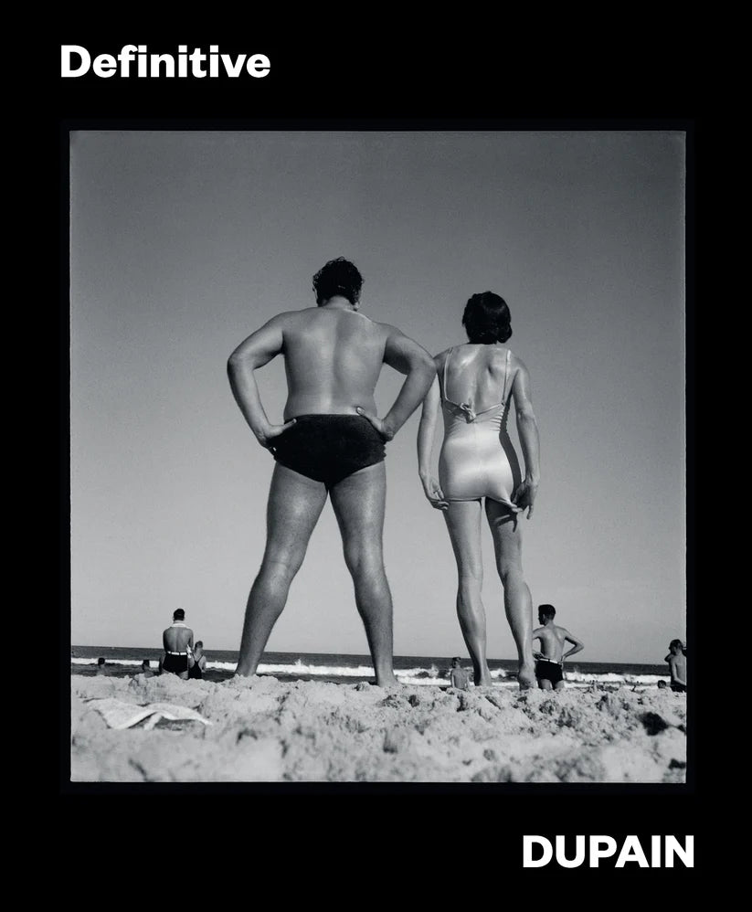 A black-and-white photographic print titled 'Definitive' by Dupain, featuring a beach scene with two people standing from behind on sand, framed by a black border with the title at top left and 'DUPAIN' at bottom right.